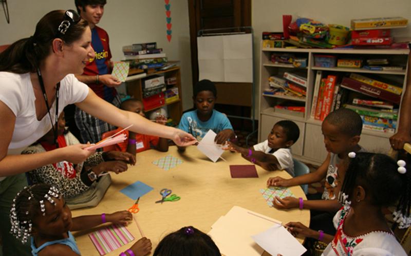 a teacher hands out craft supplies to a group of young students sitting at a table 